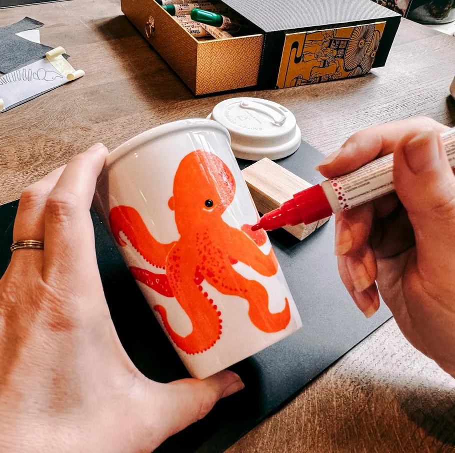 Hands drawing red octopus on white cup.