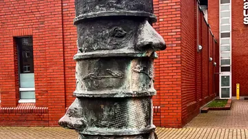Bronzeskulptur mit abstrakten Gesichtern vor Backsteingebäude.