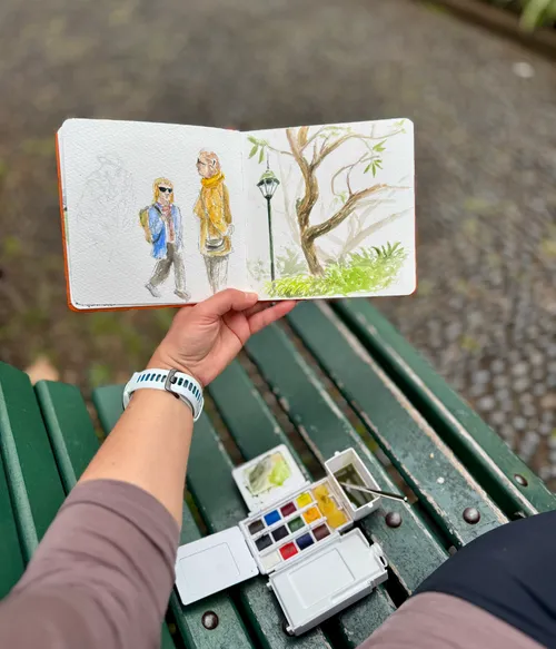 Hand holding sketchbook with watercolor painting outdoors.