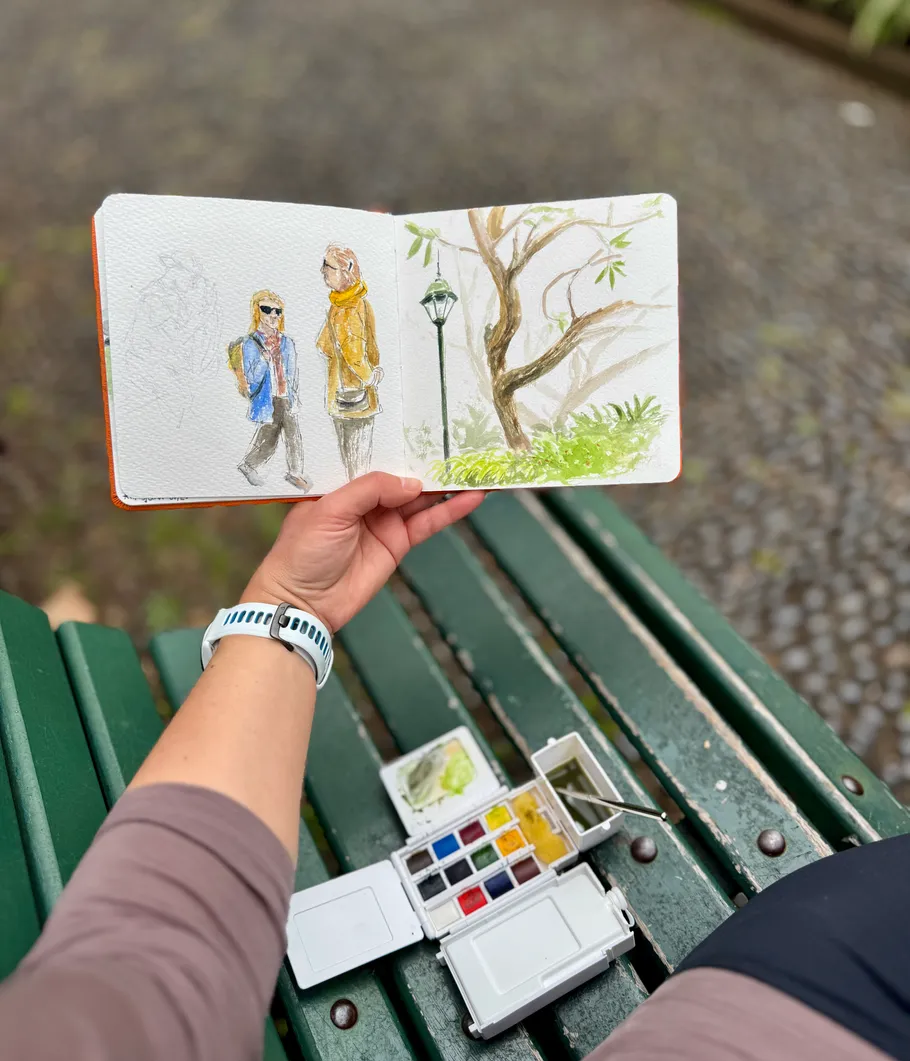 Hand holding sketchbook with watercolor painting outdoors.