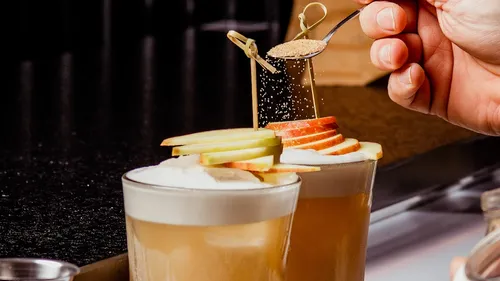 Hand adding cinnamon to apple-topped cocktails.