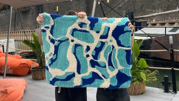 Zwei Personen halten einen blauen abstrakten Teppich im Freien.