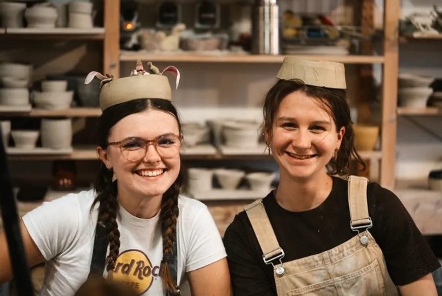 Zwei Frauen mit Tonhüten, lächelnd im Keramikstudio.