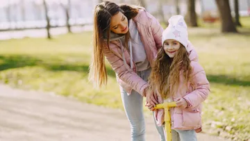 Mother helping daughter ride scooter in park.