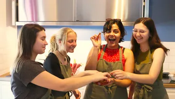 Vier Frauen in Schürzen lachen in einer Küche.