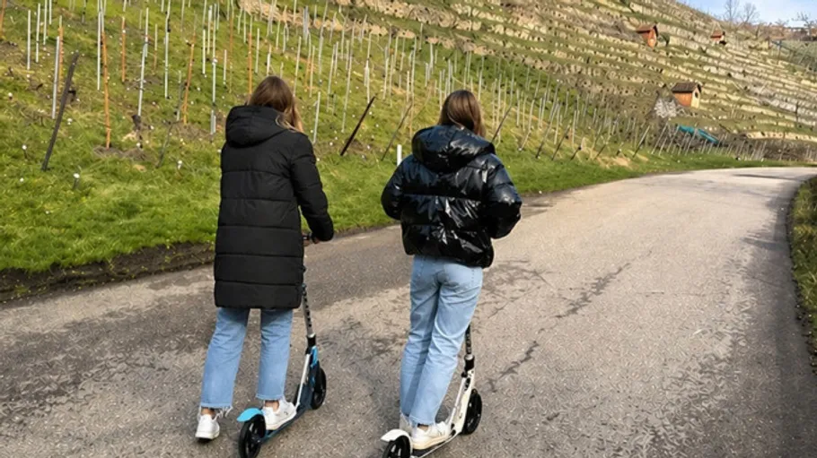 Zwei Personen fahren Roller auf einem gepflasterten Weg.