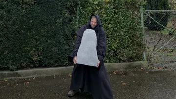 Man in cloak holding stone on wet street.