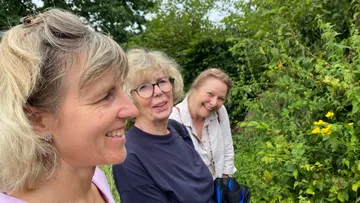 Three women smiling near bushes outdoors.