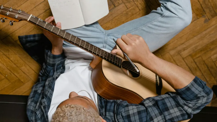 Person playing guitar, writing on notebook, wooden floor.