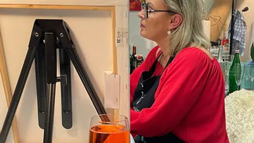Frau malt an Staffelei mit Getränk daneben.