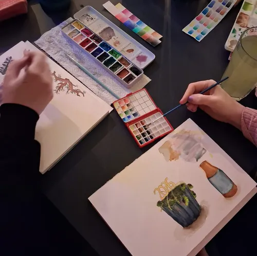 Zwei Personen malen mit Aquarellsets auf Tisch.