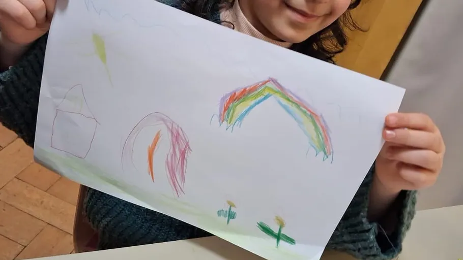 Kind hält Zeichnung mit Regenbogen und Haus.