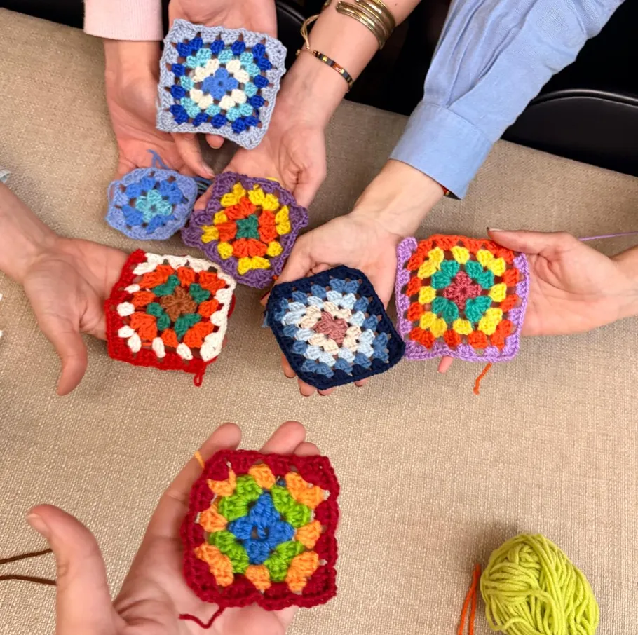 Hände halten bunte Häkelquadrate auf einem Tisch.