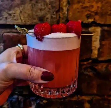 Hand hält Himbeercocktail vor Ziegelwand.