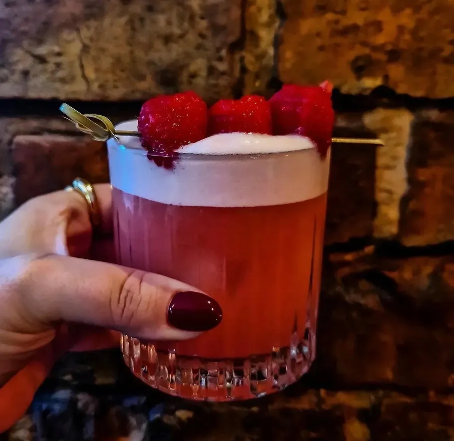 Hand holds a raspberry cocktail near brick wall.