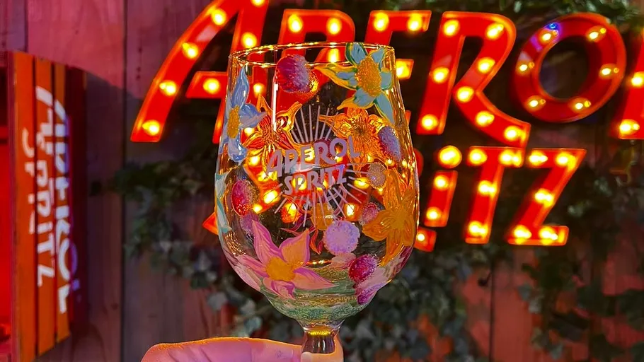 Decorated Aperol glass held, lighted sign background.