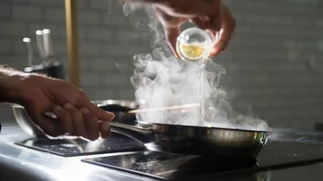 Person pours liquid into steaming pan on stove.