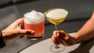 Zwei Hände halten Cocktails in einer Bar.