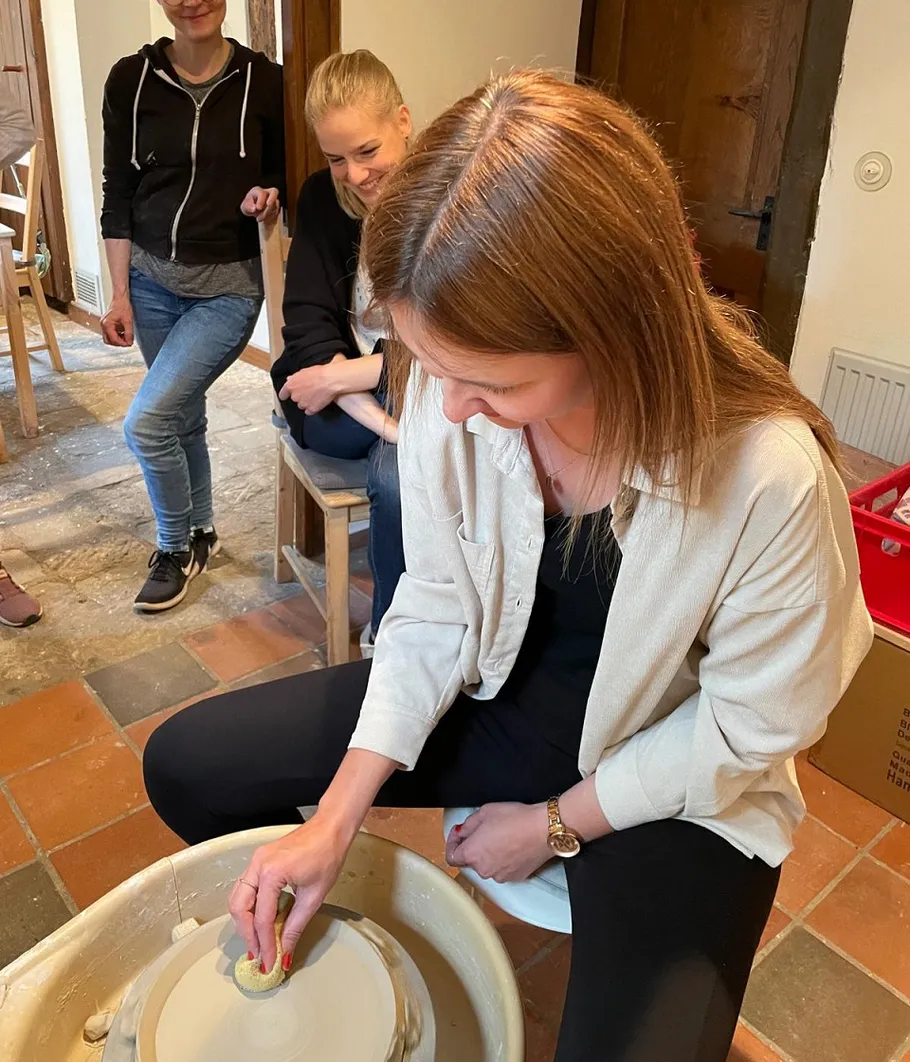 Woman sculpting clay on pottery wheel, others watching.
