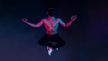 Shirtless man with mask levitating in colorful light.