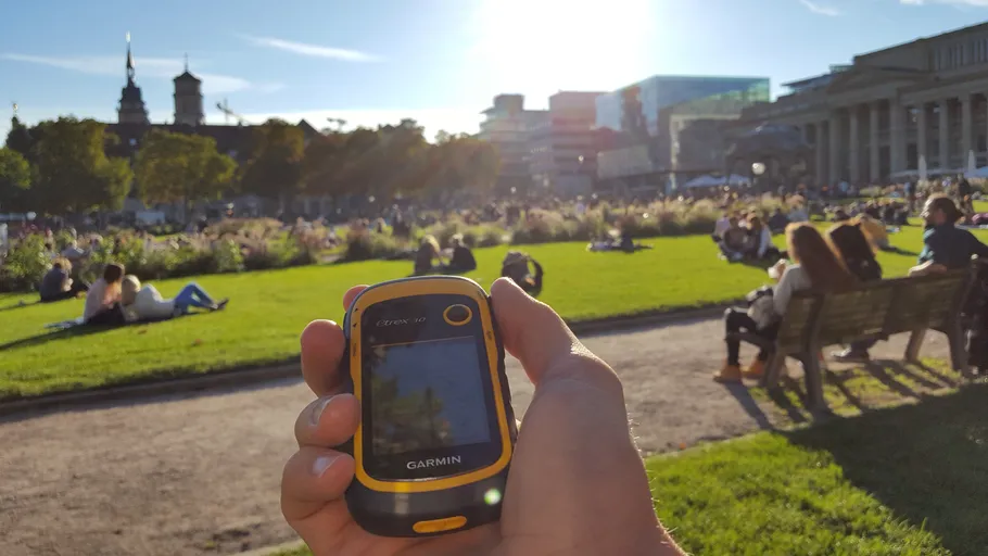 Hand hält GPS-Gerät, Park mit Menschen beim Sonnenbaden.