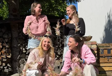 Vier Frauen lachen, halten Blumenkränze im Freien.