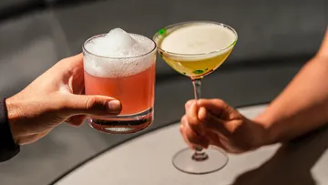 Two hands holding cocktails in a bar.
