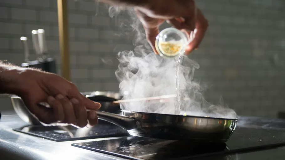 Hands adding ingredients to steaming pan on stove.