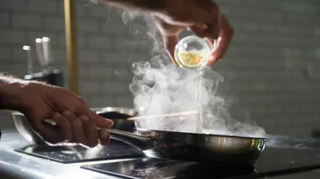 Person adding spices to steaming pan on stove.