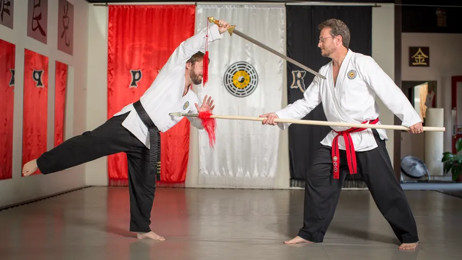 Two martial artists spar with sword and staff indoors.