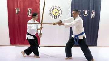 Two martial artists practicing with bamboo sticks indoors.