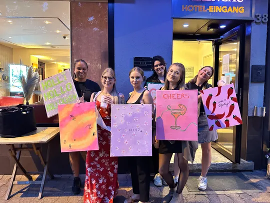 Gruppe von sechs Frauen mit bunten Gemälden draußen.