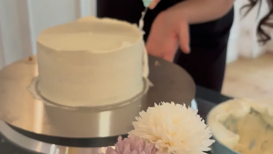 Person frosting cake on turntable, flowers nearby.