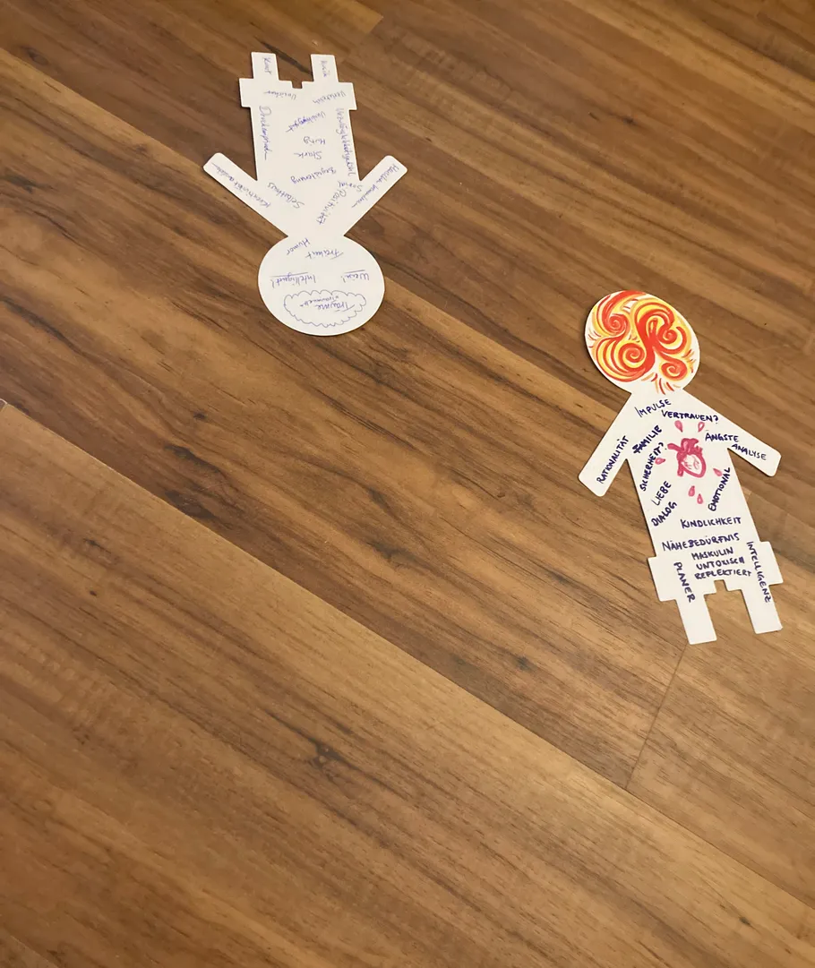 Zwei Papierfiguren mit Text auf Holzboden.