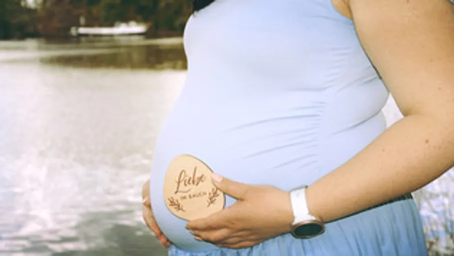 Pregnant woman holds wooden heart near lake.