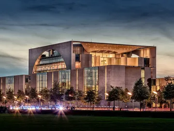 Modern government buildings glow in the evening, surrounded by trees.