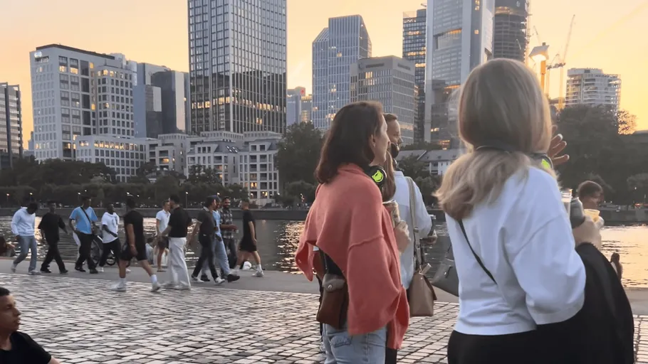 People walking by river, city skyline backdrop.