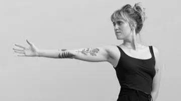Woman extends arm, displaying tattoos, neutral background.