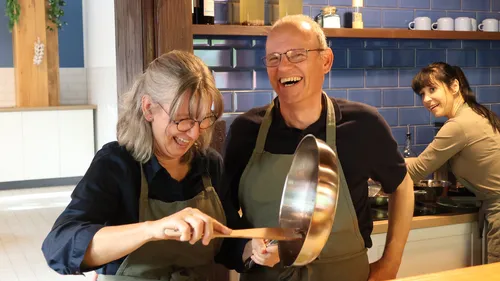 Two people smiling, holding a pan, indoor kitchen.
