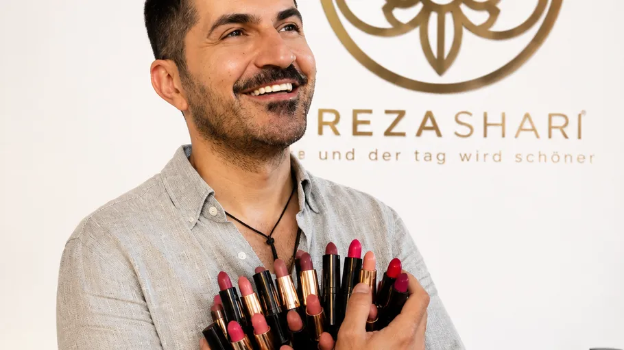 Mann hält mehrere Lippenstifte, lächelt vor Logo.