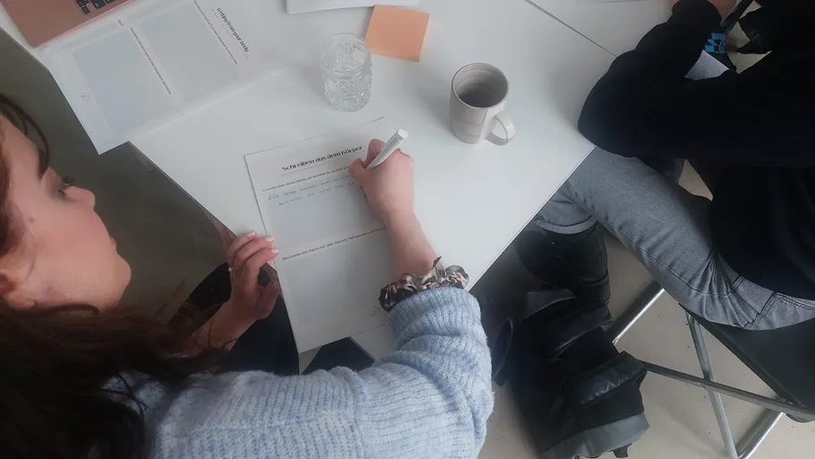 Person schreibt auf Papier an weißem Tisch.