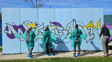 Vier Personen sprühen Graffiti auf blaue Wand.