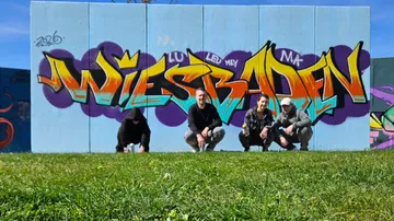 Four people crouching in front of Wiesbaden graffiti.