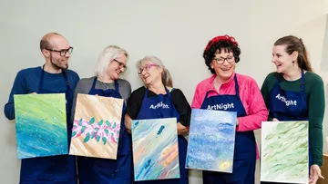 Personen halten Gemälde, lächeln, tragen blaue Schürzen.