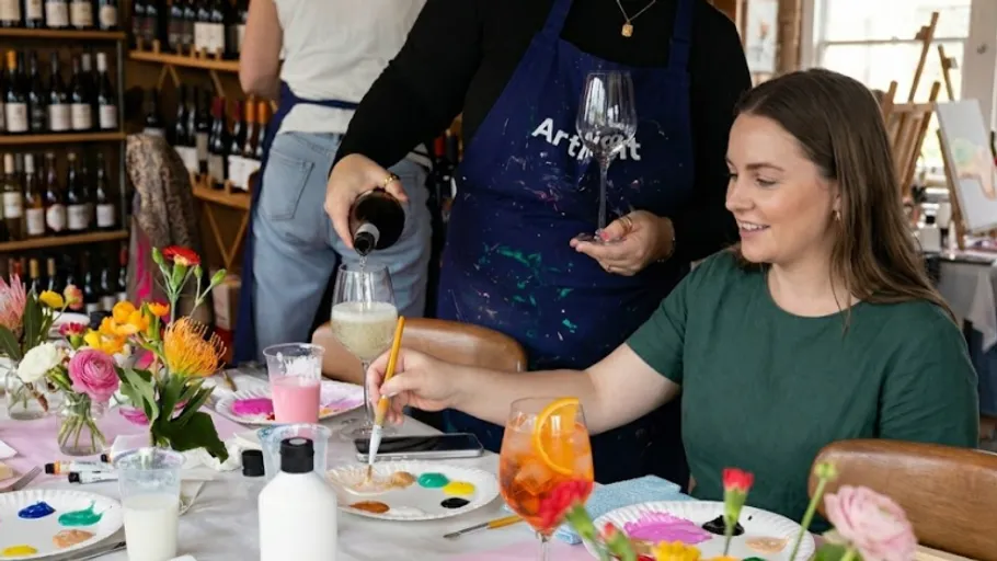 Woman painting and another pouring wine at table.