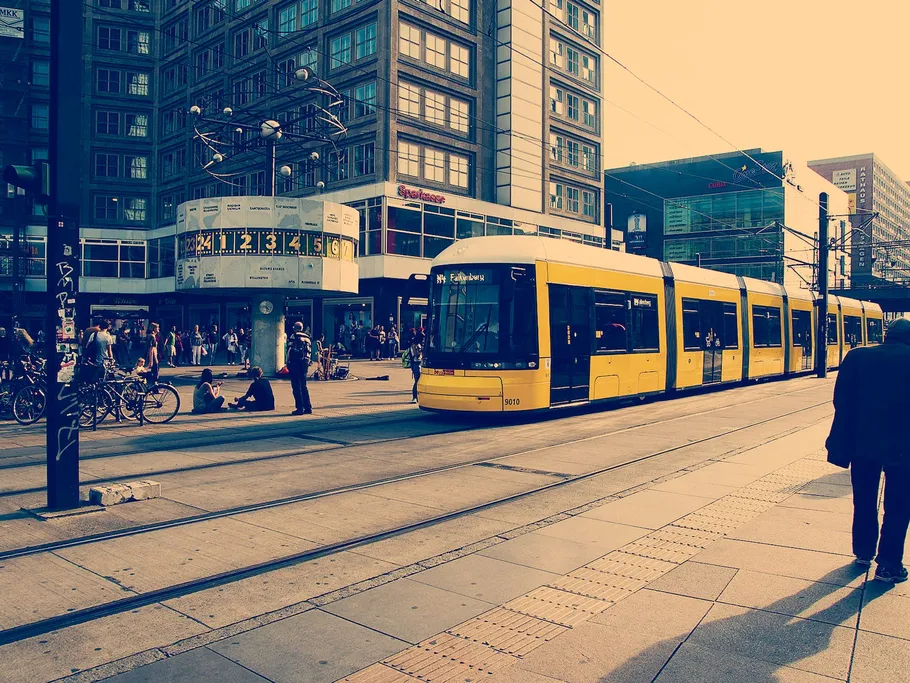 Gelbe Straßenbahn fährt durch belebten Stadtplatz, Menschen warten.