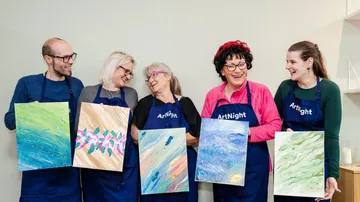 Five people smiling, holding painted canvases indoors.