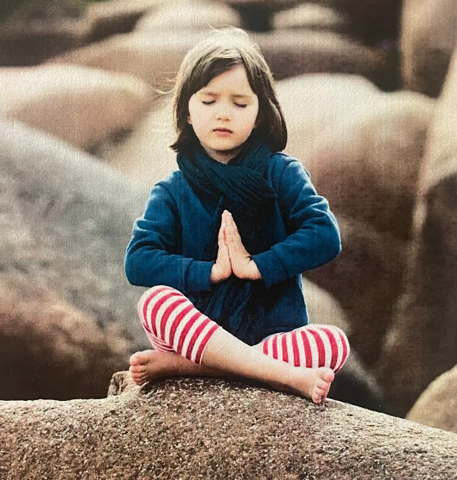 Child meditating on rocks with folded hands.