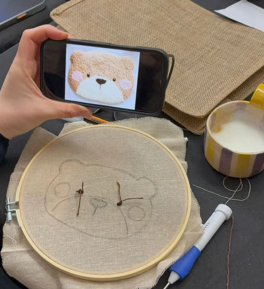 Person hält Handy mit Stickmuster, Stoff in der Nähe.