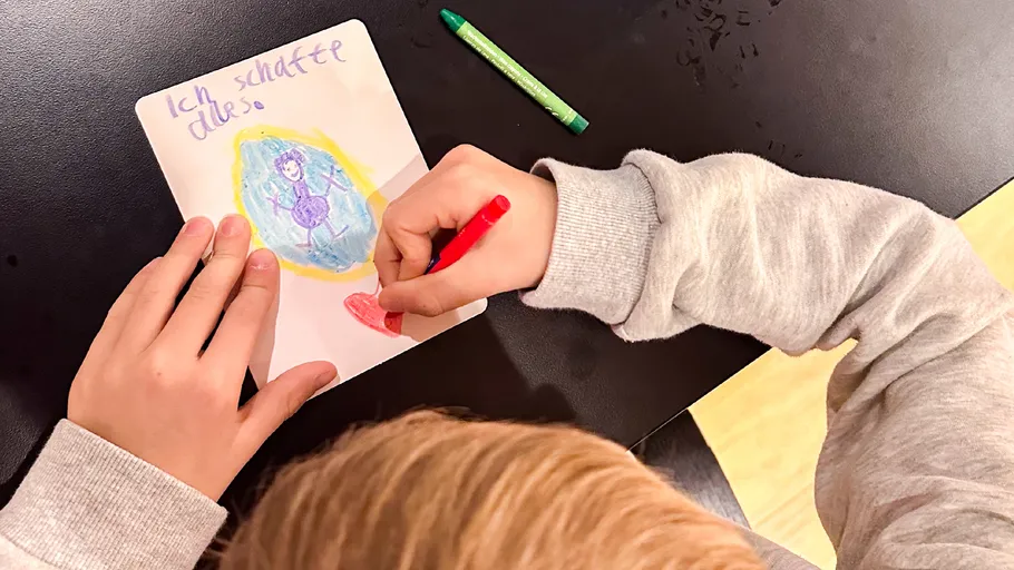 Child drawing on paper with crayons, table surface.